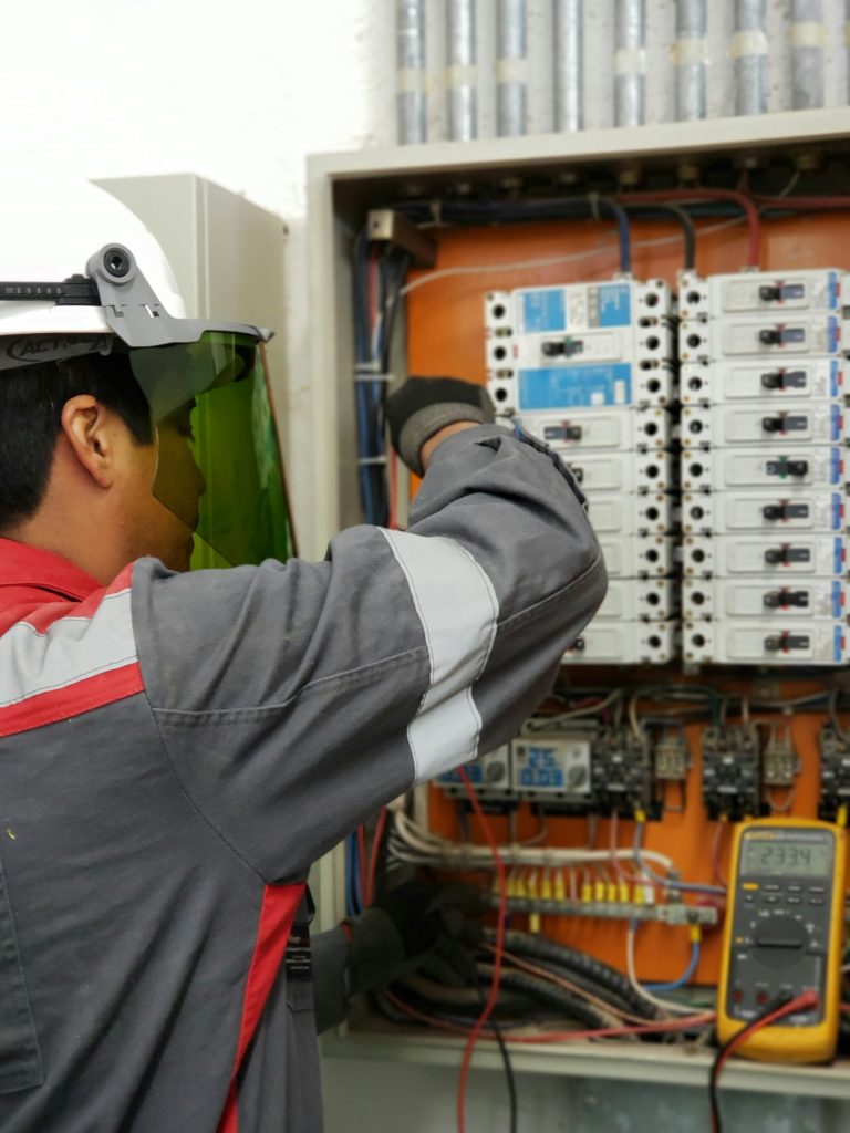 Electrician in a hard hat and face shield testing a live electrical panel