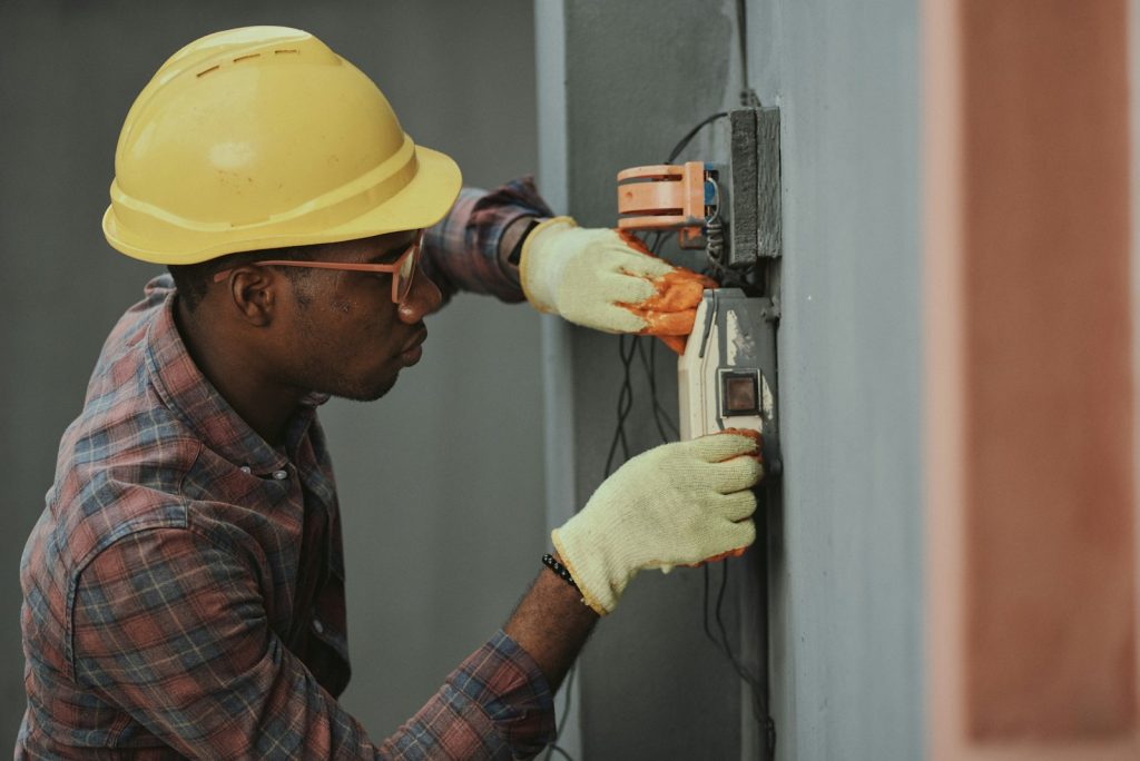 Electrician in a yellow hard hat and gloves installing wiring on a wall panel